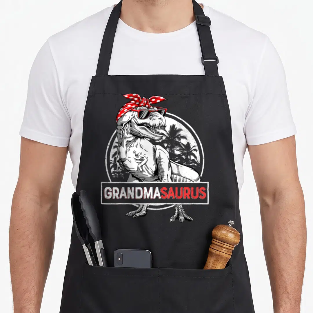 Grandmasaurus mother's apron - Cartoon T-Rex in red bandana and sunglasses with GRANDMASAURUS text banner — shown on a black cotton canvas apron against a white background