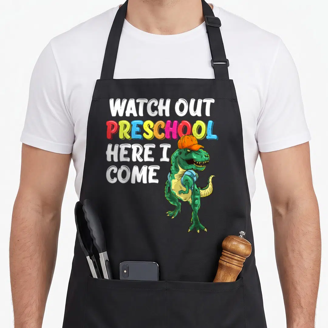 Come dinosaur apron - Stacked text Watch Out Preschool Here I Come next to a green T-Rex wearing an orange cap and blue — shown on a black cotton canvas apron against a white background