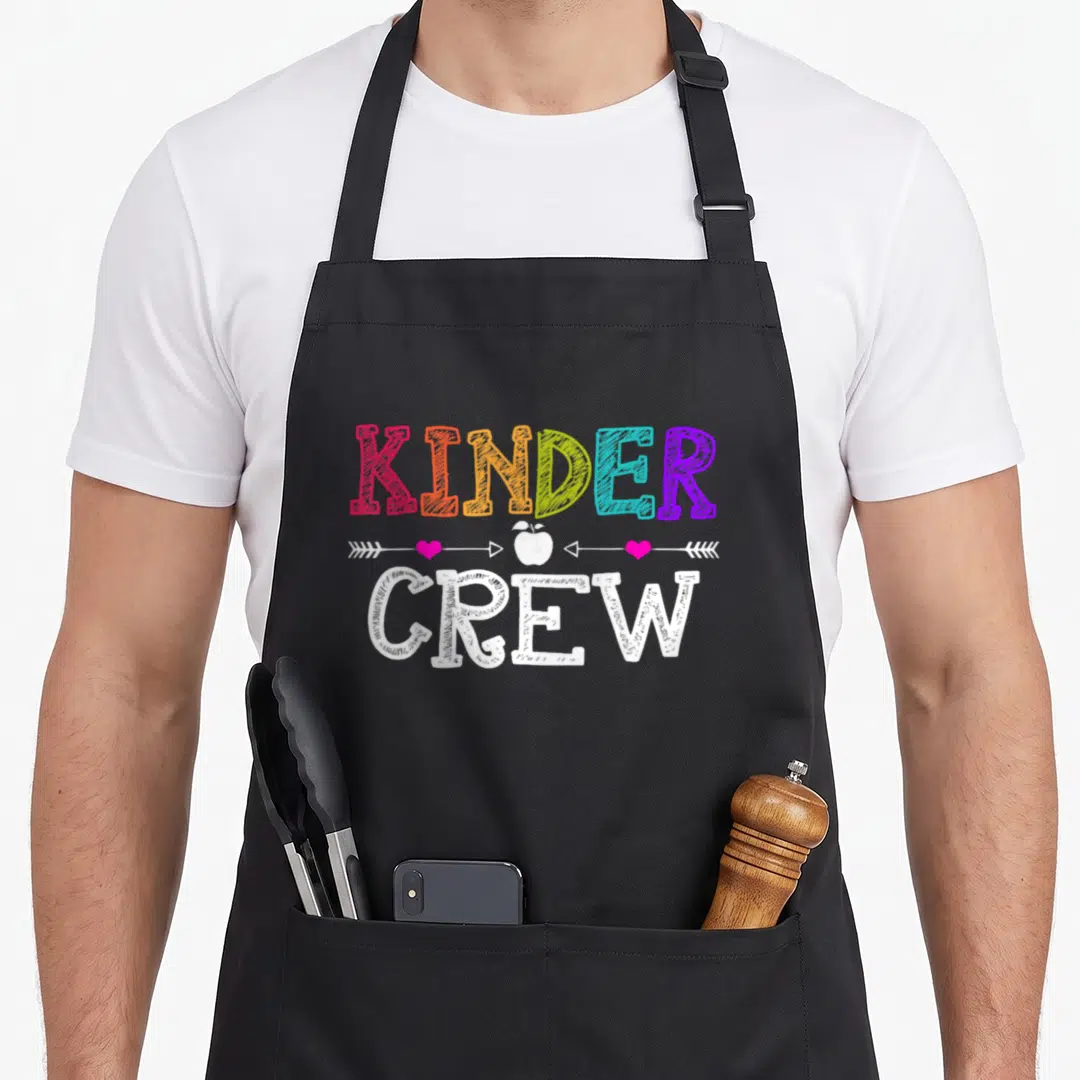 Crew kindergarten teacher apron - Rainbow KINDER text, white apple, two pink hearts, white CREW text — shown on a black cotton canvas apron against a white background