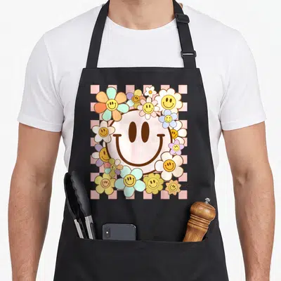 Daisy flower face on apron - Large pink smiley on pink grey checkerboard surrounded by colorful smiley-faced daisies — shown on a black cotton canvas apron against a white background