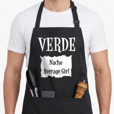 Verde taco apron - White torn paper graphic with VERDE Nacho Average Girl text in black typography — shown on a black cotton canvas apron against a white background