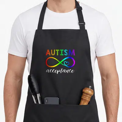 Instead autism acceptance apron - Rainbow AUTISM acceptance text with infinity symbol containing a small heart — shown on a black cotton canvas apron against a white background