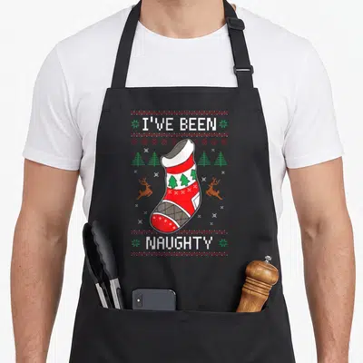 Naughty womens christmas aprons - Pixel art empty red stocking with I've Been Naughty text and reindeer border — shown on a black cotton canvas apron against a white background