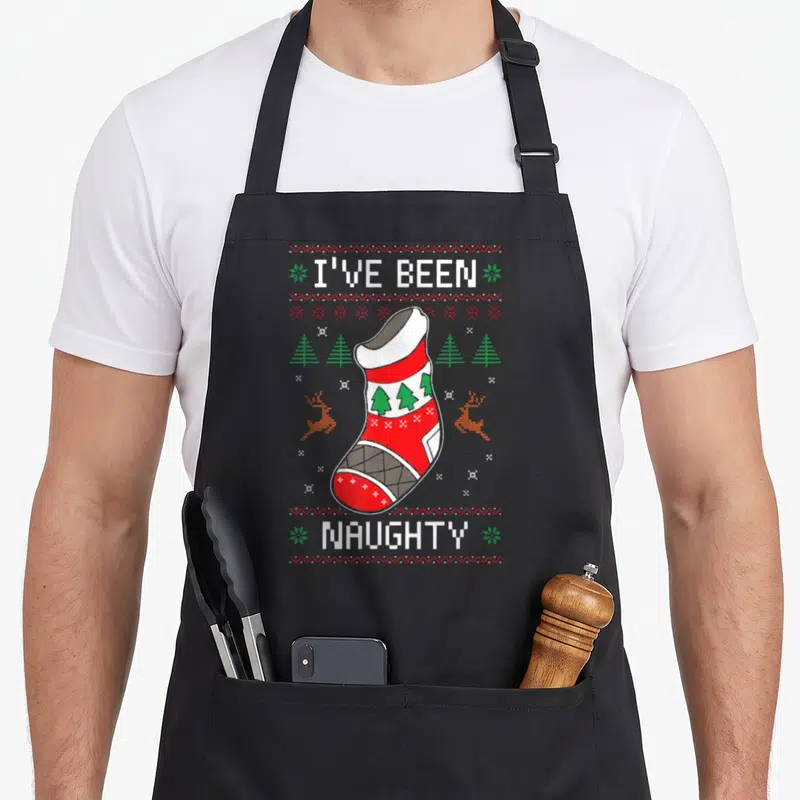 Naughty womens christmas aprons - Pixel art empty red stocking with I've Been Naughty text and reindeer border — shown on a black cotton canvas apron against a white background