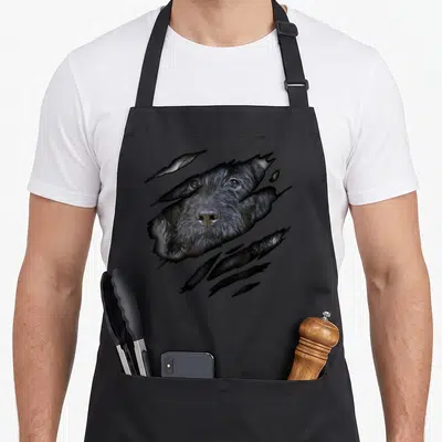 Dogdesign dog apron - Photo of a black dog face visible through a jagged ripped paper tear in grey and white — shown on a black cotton canvas apron against a white background