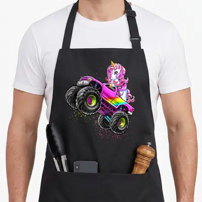 Unicorn truck apron - Cartoon unicorn leaning over pink monster truck with rainbow stripes and colorful paint splatters — shown on a black cotton canvas apron against a white background