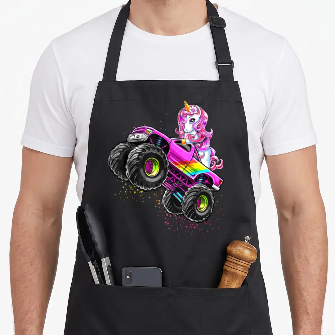 Unicorn truck apron - Cartoon unicorn leaning over pink monster truck with rainbow stripes and colorful paint splatters — shown on a black cotton canvas apron against a white background