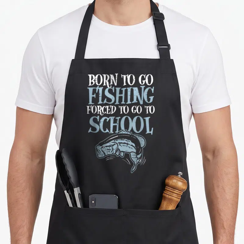 Forced fishing aprons - Distressed text BORN TO GO FISHING FORCED TO GO TO SCHOOL above light blue bass fish drawing — shown on a black cotton canvas apron against a white background