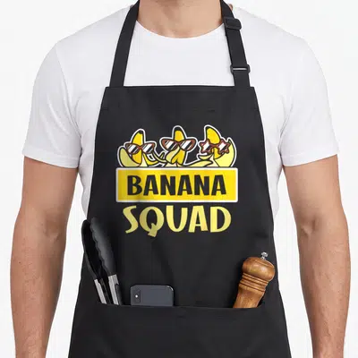 Squad bananas banana apron - Three cartoon bananas with sunglasses above yellow banner reading BANANA SQUAD — shown on a black cotton canvas apron against a white background