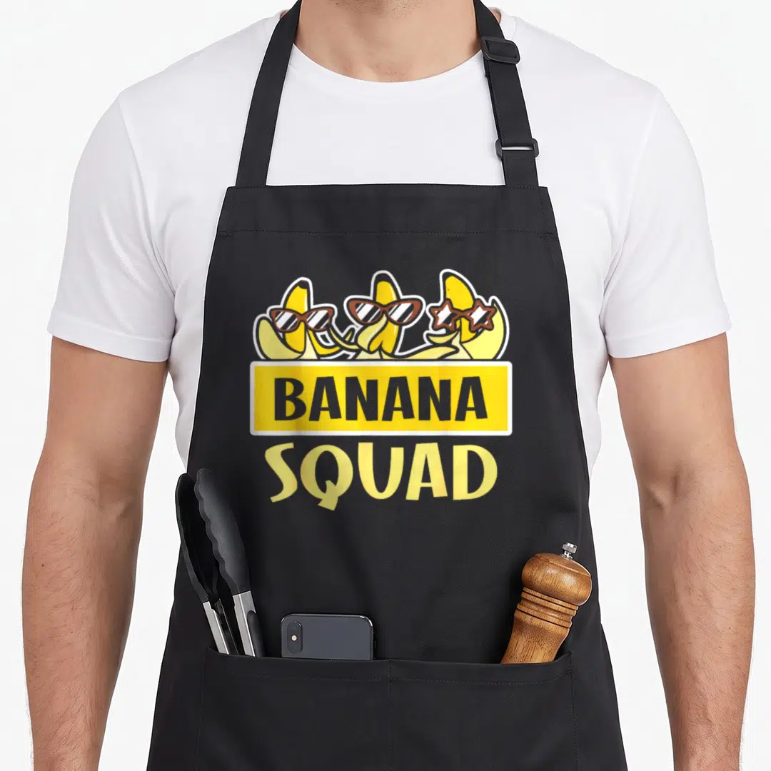 Squad bananas banana apron - Three cartoon bananas with sunglasses above yellow banner reading BANANA SQUAD — shown on a black cotton canvas apron against a white background