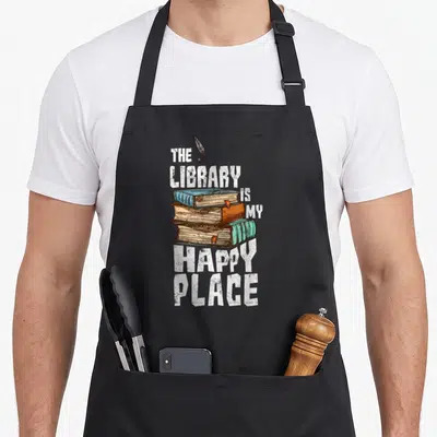 Librarian book apron - Stack of three blue, brown, and green books with inkwell and distressed text — shown on a black cotton canvas apron against a white background
