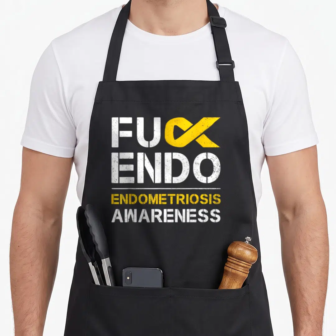 Endo endometriosis ribbon apron - White and yellow stacked text with yellow awareness ribbon replacing K in FUCK — shown on a black cotton canvas apron against a white background