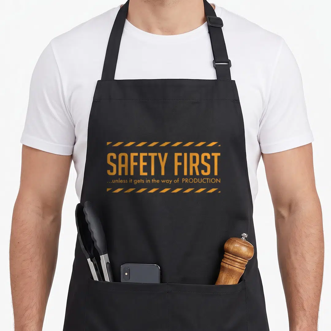 Unless safety apron - Orange typography reading Safety First unless it gets in the way of production between striped — shown on a black cotton canvas apron against a white background