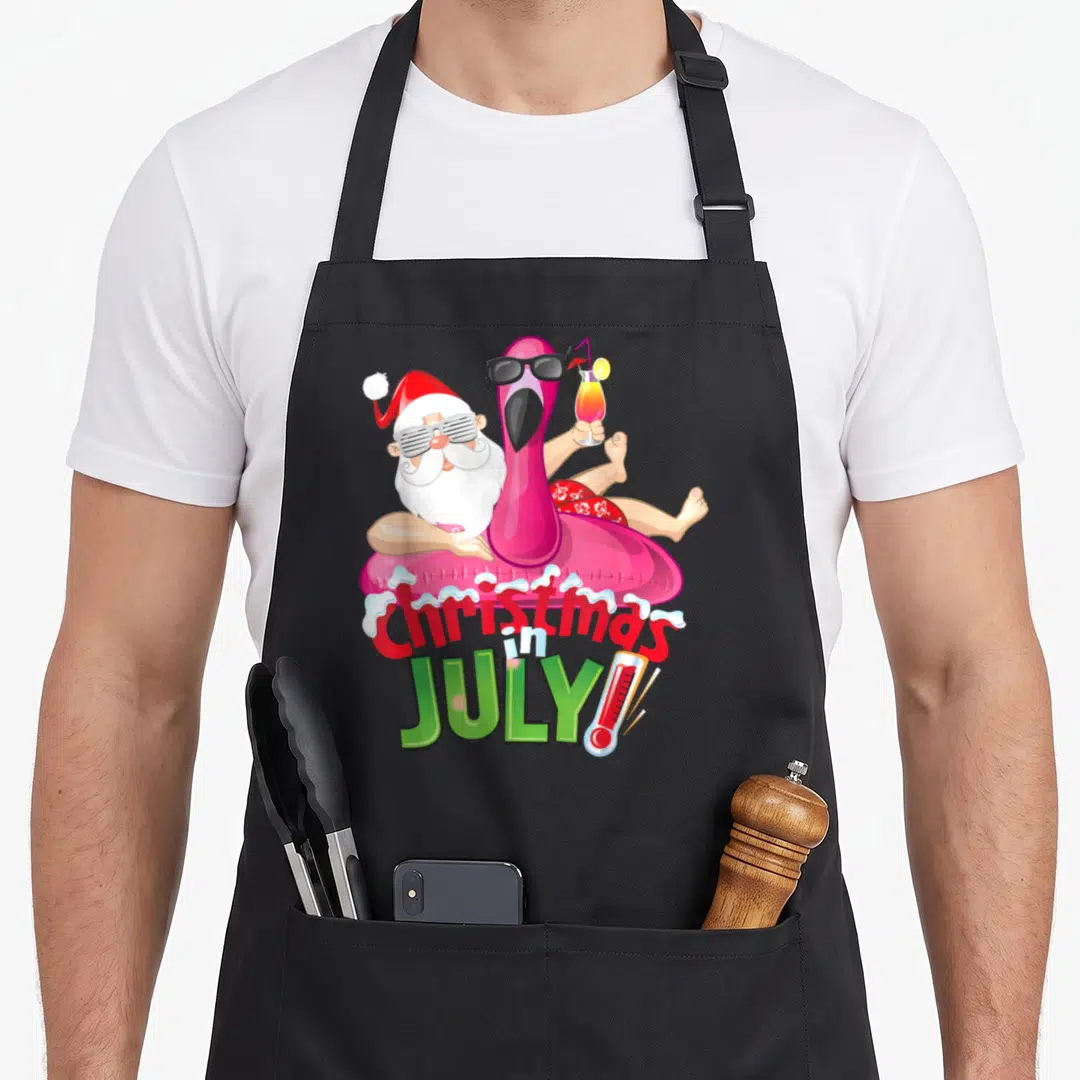 July christmas flamingo apron - Santa and pink flamingo in float with Christmas in July text and thermometer — shown on a black cotton canvas apron against a white background