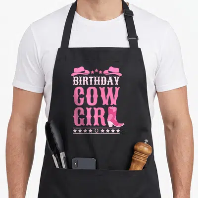 Western cowgirl aprons - Pink Birthday Cow Girl text with cowboy hats, stars, horseshoe, and boot — shown on a black cotton canvas apron against a white background