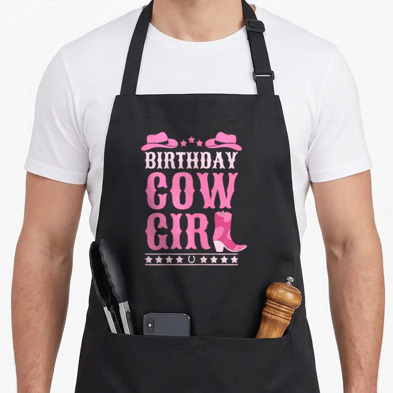 Western cowgirl aprons - Pink Birthday Cow Girl text with cowboy hats, stars, horseshoe, and boot — shown on a black cotton canvas apron against a white background