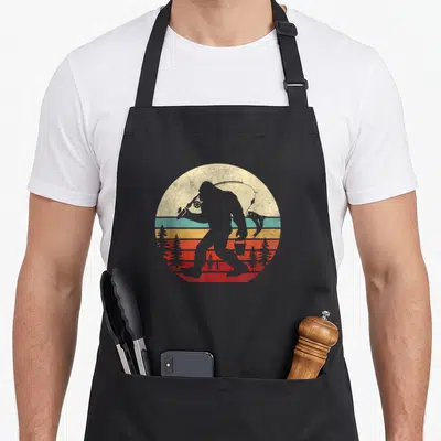 Bigfoot fishing apron - Grey bigfoot silhouette holding fishing rod and bucket against retro striped sun with red pine — shown on a black cotton canvas apron against a white background