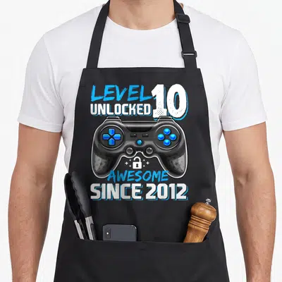 10th birthday age video game apron - Black game controller with blue buttons and LEVEL UNLOCKED 10 text — shown on a black cotton canvas apron against a white background