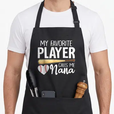 Baseball player grandma apron - Stacked text My Favorite Player Calls Me Nana with tan bat and red stitched heart — shown on a black cotton canvas apron against a white background