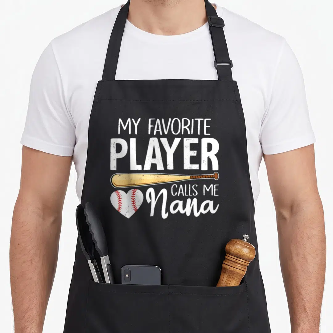Baseball player grandma apron - Stacked text My Favorite Player Calls Me Nana with tan bat and red stitched heart — shown on a black cotton canvas apron against a white background