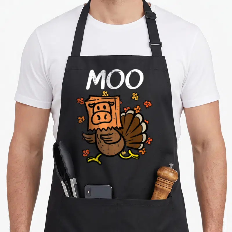 Cow thanksgiving apron - Cartoon turkey wearing orange cow paper bag mask with MOO text and fall leaves — shown on a black cotton canvas apron against a white background