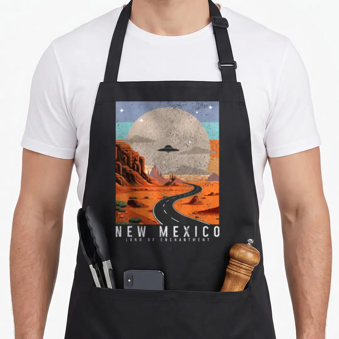 Pride ufo mexico apron - Retro desert landscape with UFO, winding road, large sun, and NEW MEXICO text — shown on a black cotton canvas apron against a white background