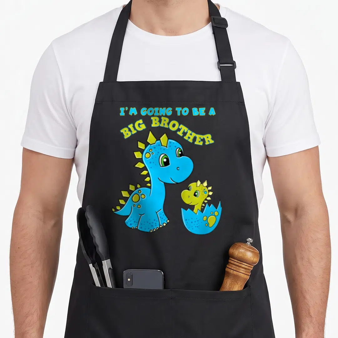 Brother dinosaur apron - Blue and yellow cartoon dinosaurs with I'M GOING TO BE A BIG BROTHER text — shown on a black cotton canvas apron against a white background