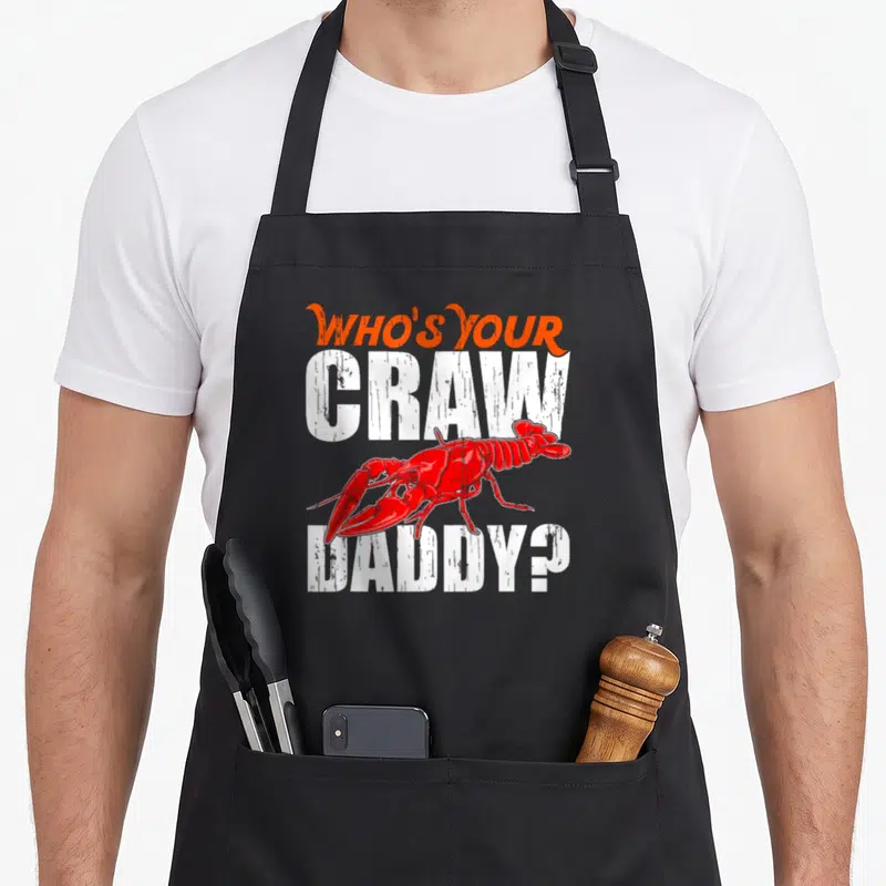 Cajun crawfish apron - Distressed orange and white text reading Who's Your Craw Daddy with red crawfish between lines — shown on a black cotton canvas apron against a white background