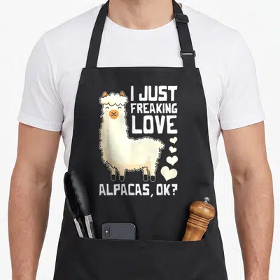 Freaking alpaca apron - Cartoon alpaca in cream and tan with stacked text and four hearts — shown on a black cotton canvas apron against a white background