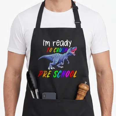 1st pre dinosaur apron - Blue T-Rex cartoon between white and rainbow text reading I'm ready TO CRUSH PRE SCHOOL — shown on a black cotton canvas apron against a white background