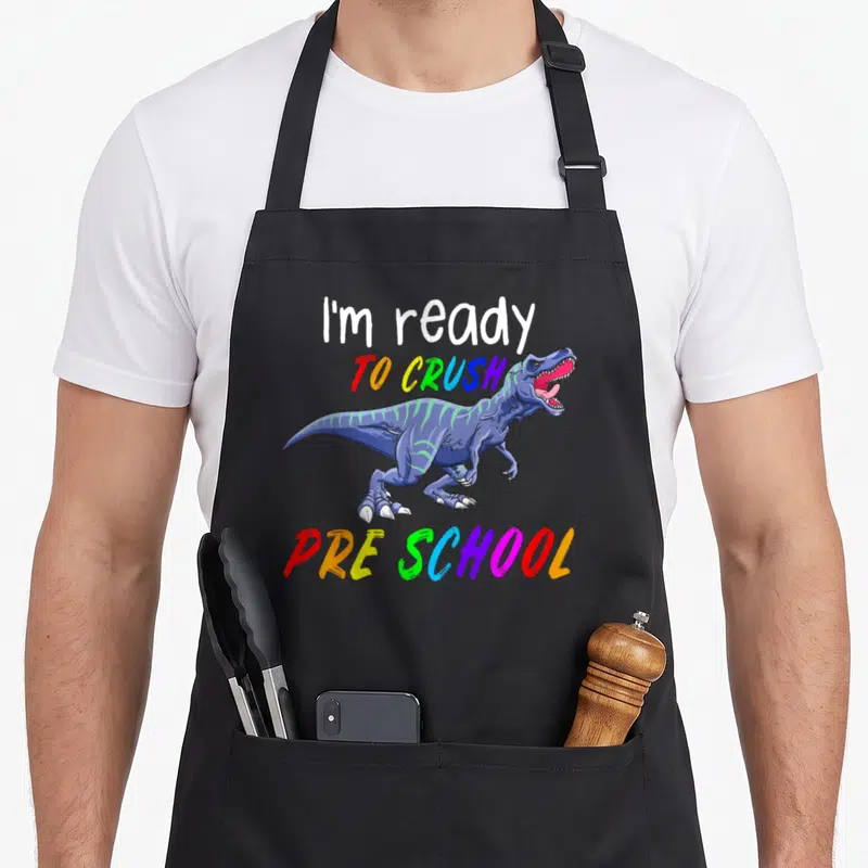 1st pre dinosaur apron - Blue T-Rex cartoon between white and rainbow text reading I'm ready TO CRUSH PRE SCHOOL — shown on a black cotton canvas apron against a white background