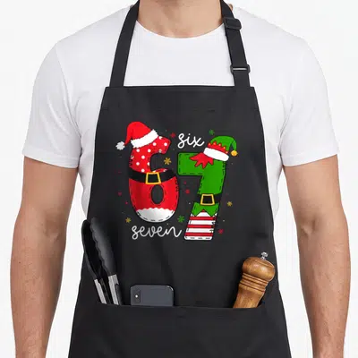 Meme six seven christmas apron - Number 67 dressed as Santa and elf with snowflakes and stars — shown on a black cotton canvas apron against a white background