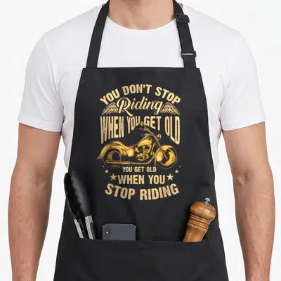 Stop cycling apron - Retro yellow-orange motorcycle with wings and stars surrounding the text YOU DON'T STOP Riding — shown on a black cotton canvas apron against a white background