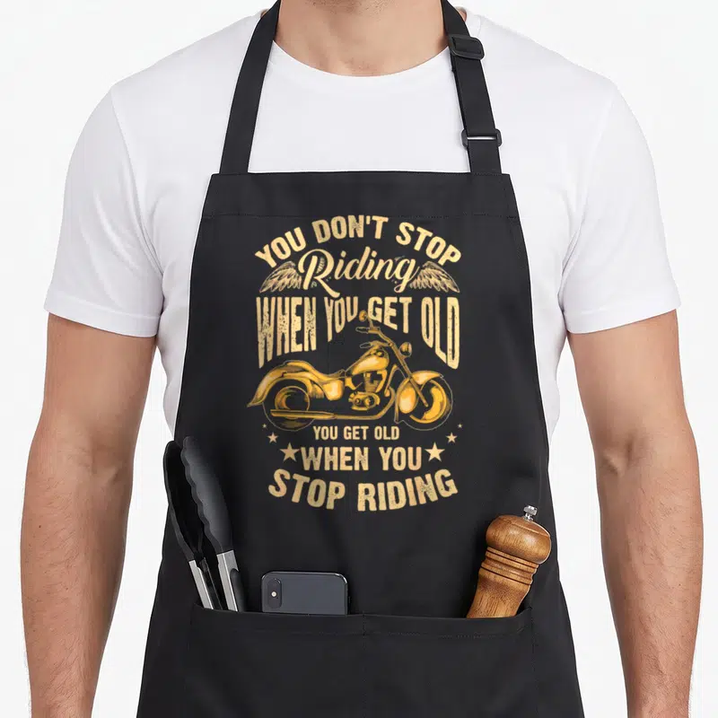 Stop cycling apron - Retro yellow-orange motorcycle with wings and stars surrounding the text YOU DON'T STOP Riding — shown on a black cotton canvas apron against a white background