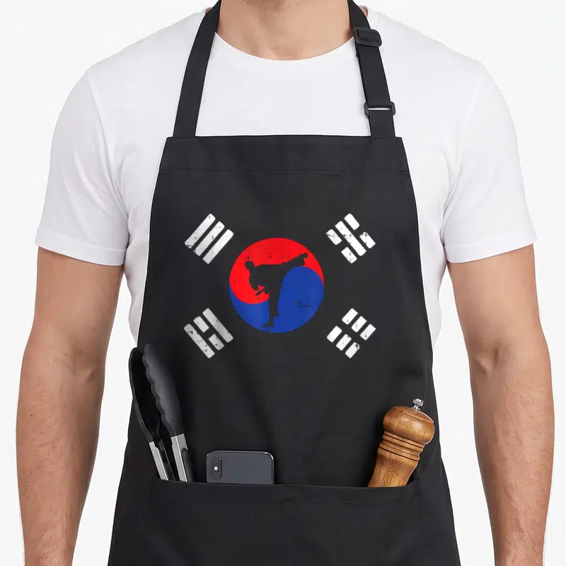Martial korean apron - Distressed red and blue Taegeuk with kicking martial artist silhouette and four white trigrams — shown on a black cotton canvas apron against a white background