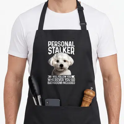 Maltese personal chef apron - White Maltese dog head with Personal Stalker text on grey background — shown on a black cotton canvas apron against a white background