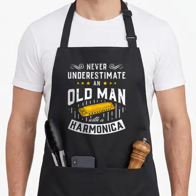 Music harmonica apron - Stacked white text never underestimate an old man with a harmonica above a yellow harmonica — shown on a black cotton canvas apron against a white background