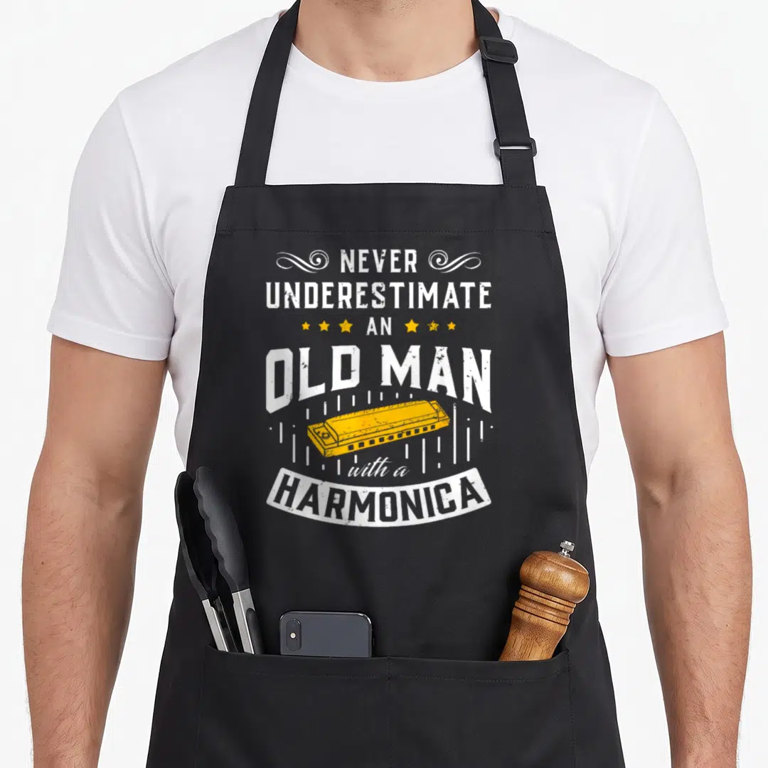 Music harmonica apron - Stacked white text never underestimate an old man with a harmonica above a yellow harmonica — shown on a black cotton canvas apron against a white background