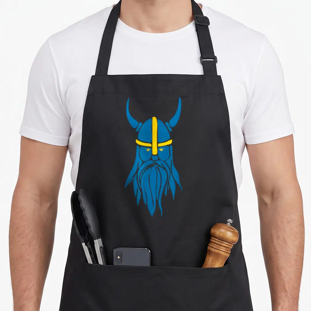 Swedish scandinavian aprons - Stylized blue and yellow viking helmet with horns and long beard on black fabric — shown on a black cotton canvas apron against a white background
