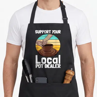 Pottery dealer apron - Retro sunset circle with hands shaping brown pot on wheel, white text reads Support Your Local — shown on a black cotton canvas apron against a white background
