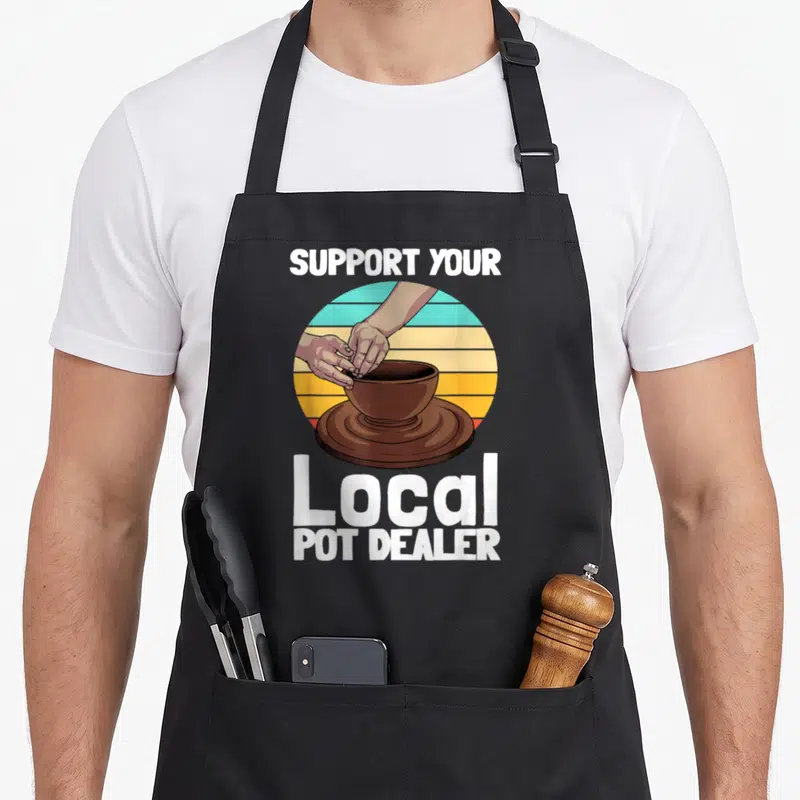 Pottery dealer apron - Retro sunset circle with hands shaping brown pot on wheel, white text reads Support Your Local — shown on a black cotton canvas apron against a white background