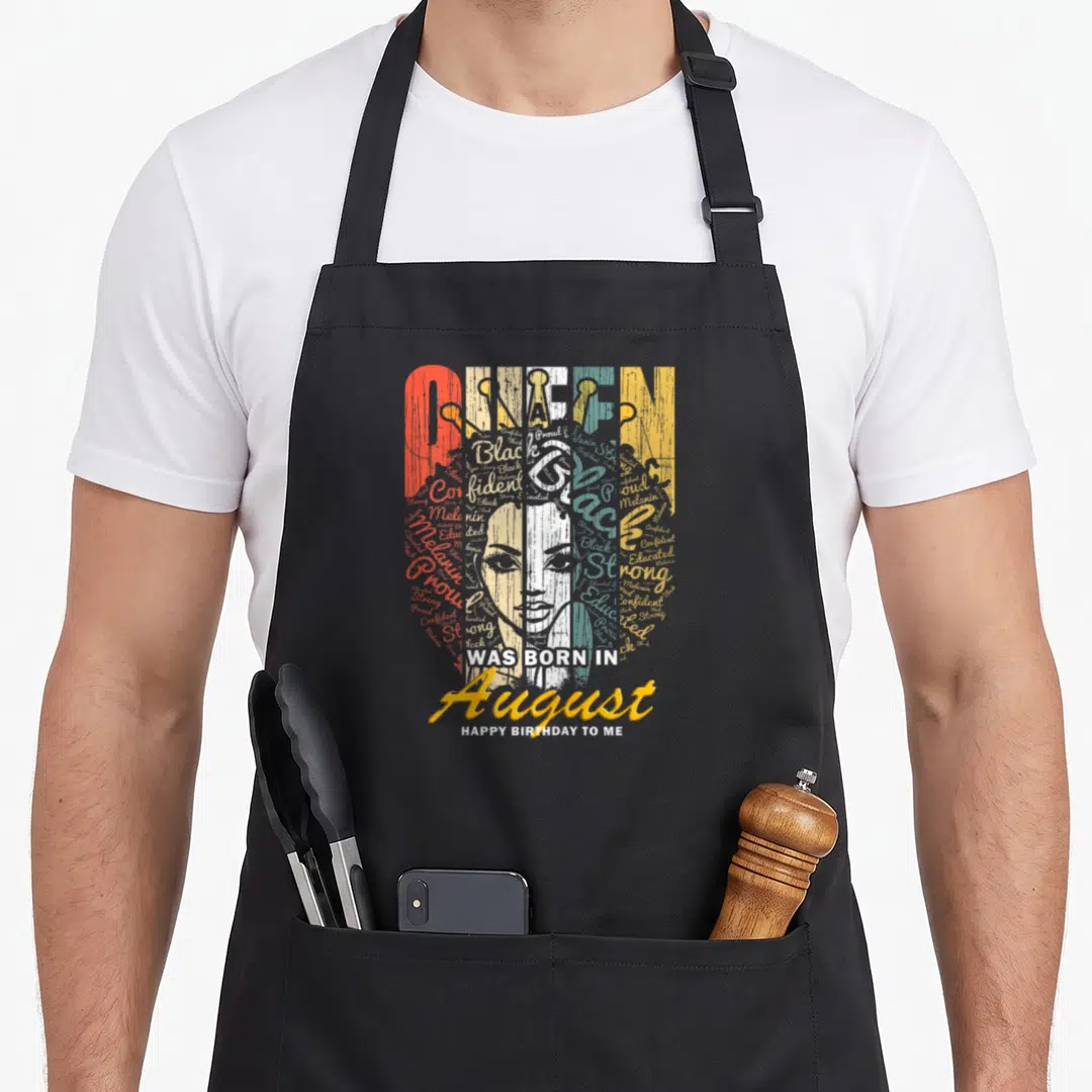 Funny august birthday apron - Distressed QUEEN graphic with woman's face and August birthday text in red white blue — shown on a black cotton canvas apron against a white background