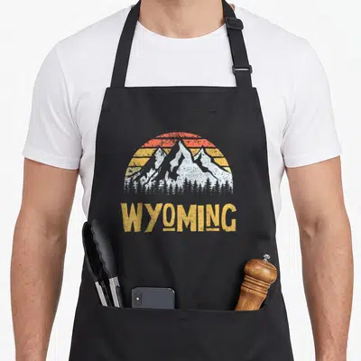 Mountain wyoming boy apron - Retro white mountains with orange and yellow striped sun and WYOMING text — shown on a black cotton canvas apron against a white background