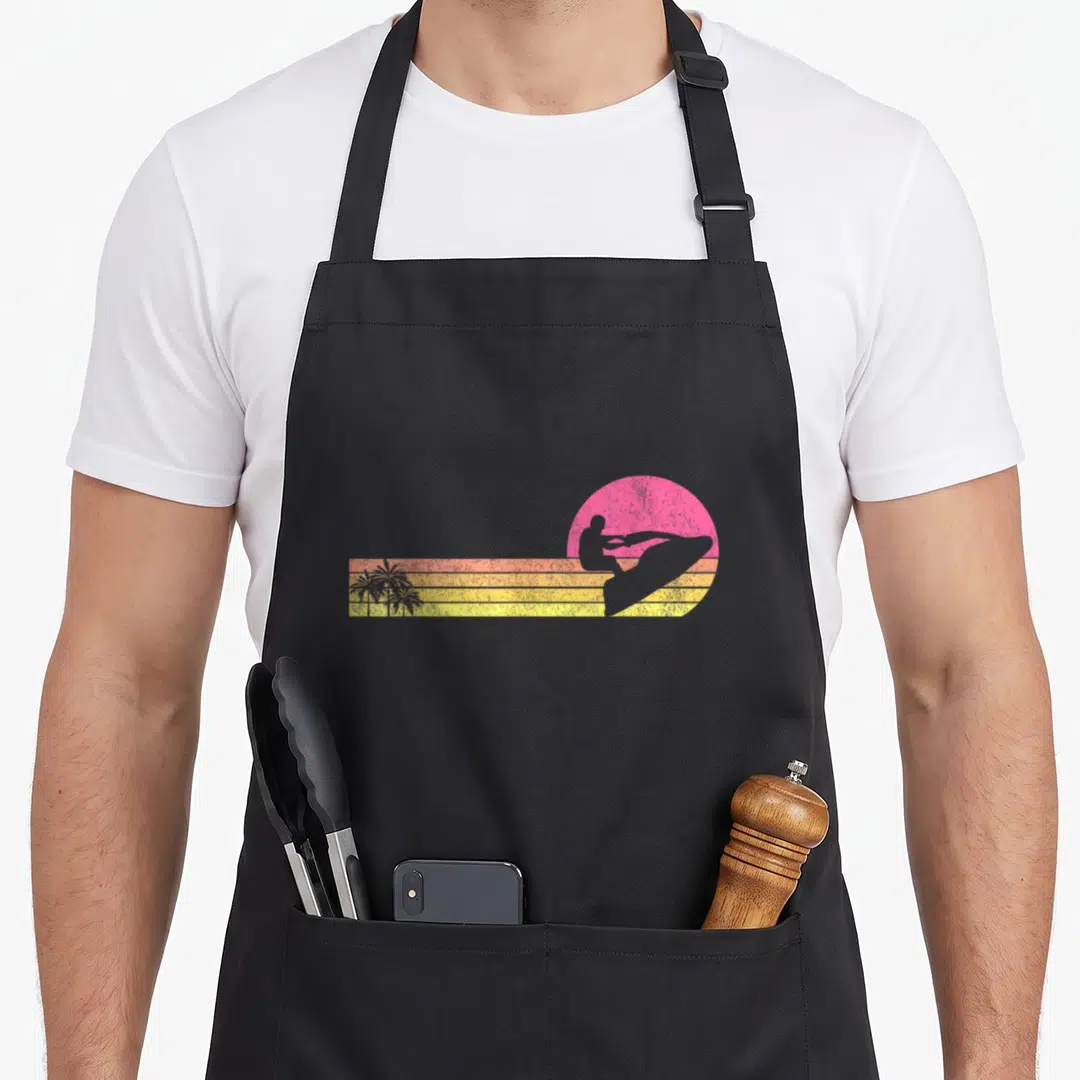 Ski skier jetski apron - Retro jet ski silhouette on pink sun with orange yellow stripes and three palm trees — shown on a black cotton canvas apron against a white background