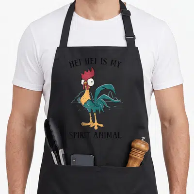 Hei animal apron - Cartoon rooster with red comb standing between HEI HEI IS MY SPIRIT ANIMAL text — shown on a black cotton canvas apron against a white background