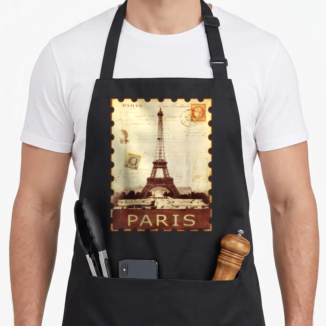 Stamp paris apron - Vintage Eiffel Tower illustration inside a brown scalloped stamp border with handwritten script — shown on a black cotton canvas apron against a white background