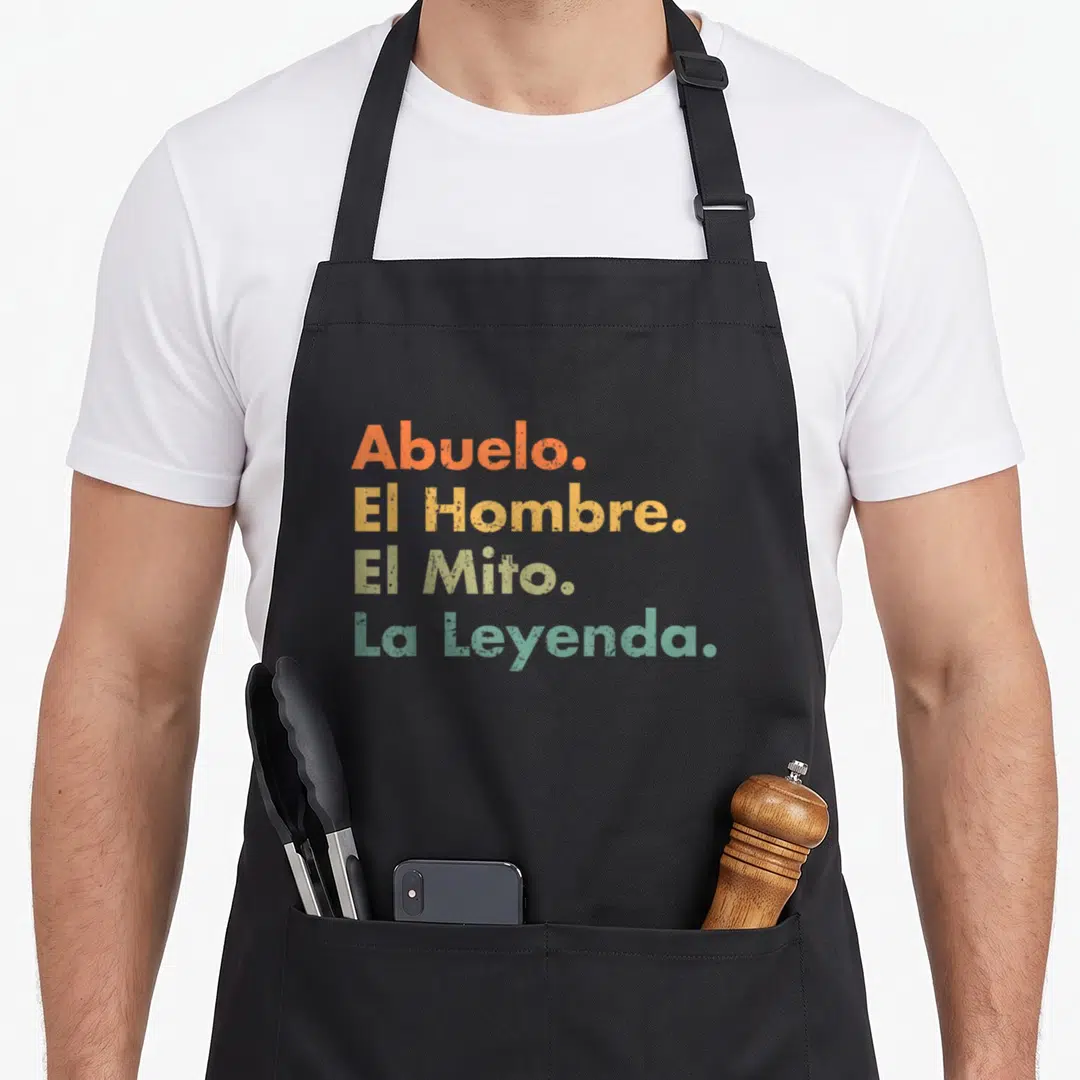Grandpa spanish aprons - Stacked Spanish text Abuelo El Hombre El Mito La Leyenda in orange yellow olive green and teal — shown on a black cotton canvas apron against a white background