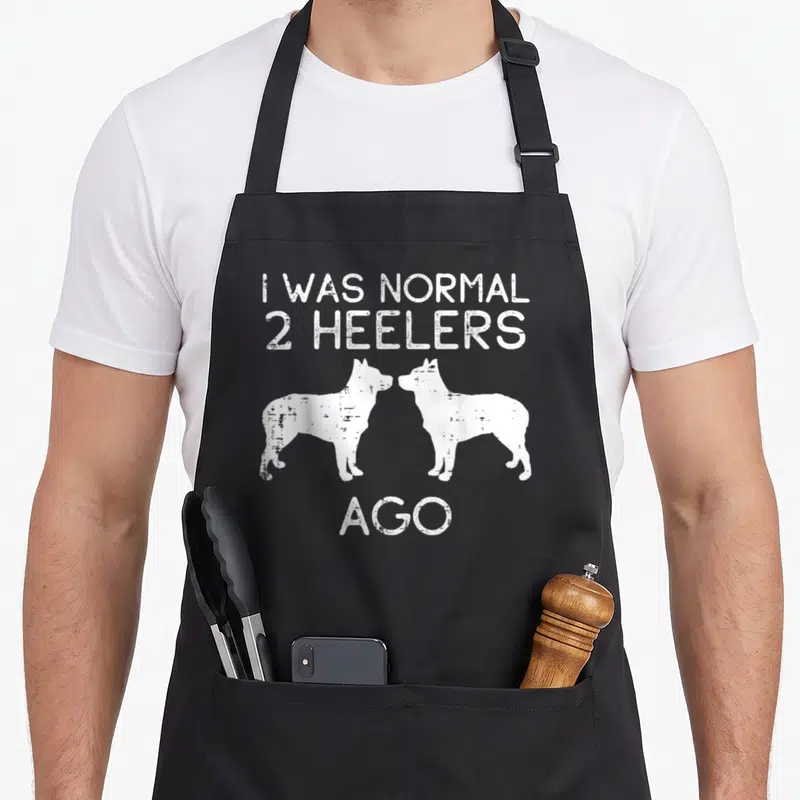 Normal dog kitchen apron - White distressed text I WAS NORMAL 2 HEELERS AGO above two facing dog silhouettes — shown on a black cotton canvas apron against a white background