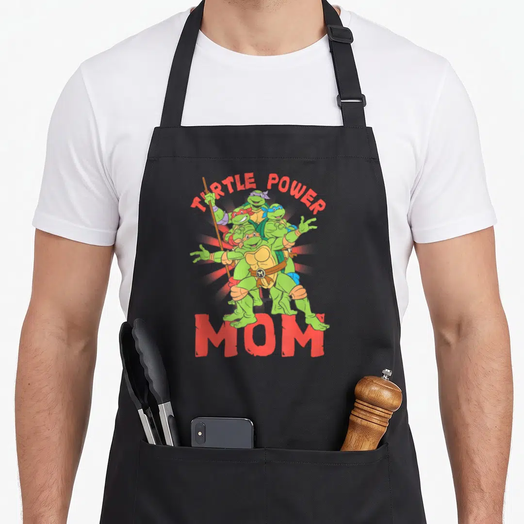 Power turtle apron - Four green turtles with colored masks under TURTLE POWER and above MOM text — shown on a black cotton canvas apron against a white background