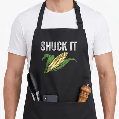 Shuck farmers market aprons - Distressed white SHUCK IT text above a yellow corn cob with green husks — shown on a black cotton canvas apron against a white background